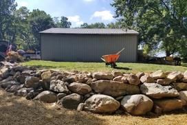 A stone retaining wall sits in front of a gray metal building on a grassy lawn with an orange wheelbarrow nearby.