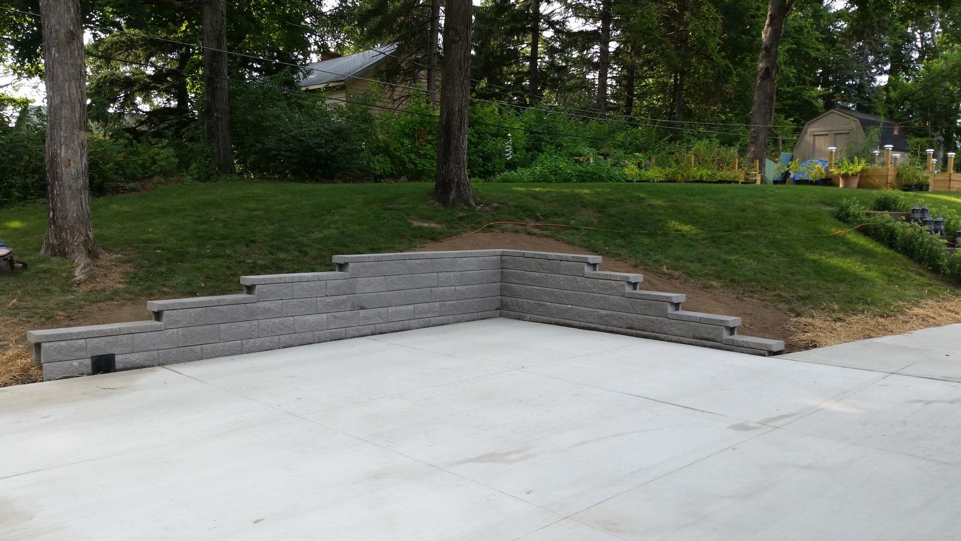 A stepped gray concrete block retaining wall borders a smooth paved patio at the base of a grassy, tree-lined hill.