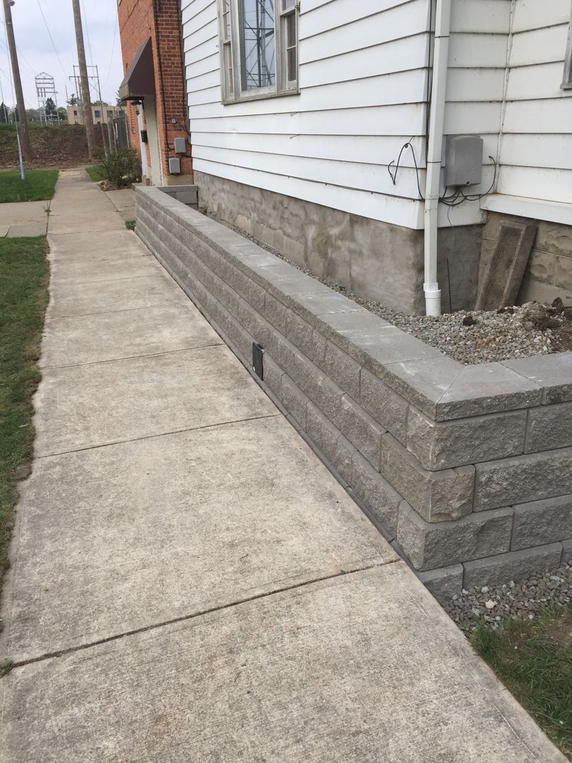 A concrete sidewalk runs alongside a grey, tiered stone retaining wall built against the side of a white house.
