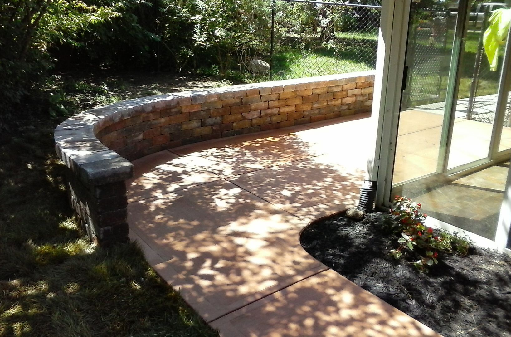 A curved, stone retaining wall borders a tan concrete patio next to a glass enclosed room with dark mulch landscaping.