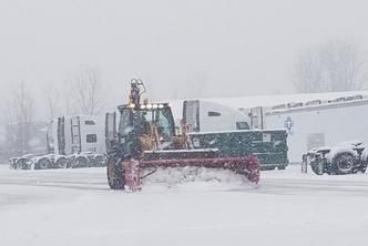 A tractor with a snowplow clears a snowy parking lot filled with parked semi-trucks during a blizzard.