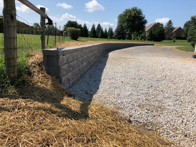 A curved grey stone retaining wall borders a gravel driveway next to a fenced grassy field under a sunny blue sky.