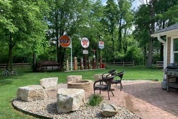 A backyard patio with a fire pit, seating, and a collection of vintage gas station signs on poles in the grassy lawn.