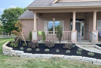 A stone retaining wall surrounds a mulched garden bed with small shrubs and a green garden tool in front of a brick house.