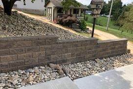 A brown stone retaining wall sits in front of a rock-covered hill next to a paved walkway and a small cottage.