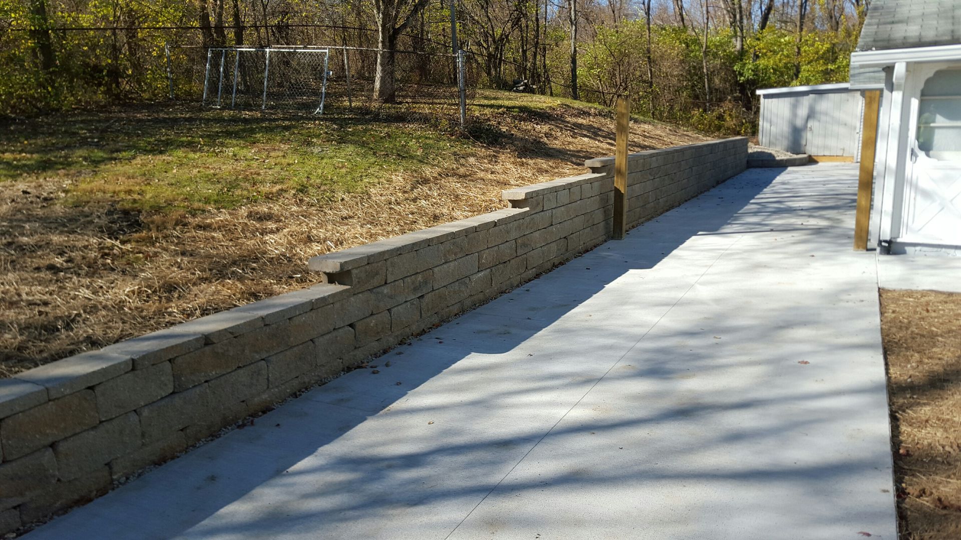 A long, beige concrete block retaining wall lines a newly poured concrete patio beside a wooded area.