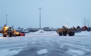 Two construction vehicles clearing snow in an open, snowy lot under a gray sky.