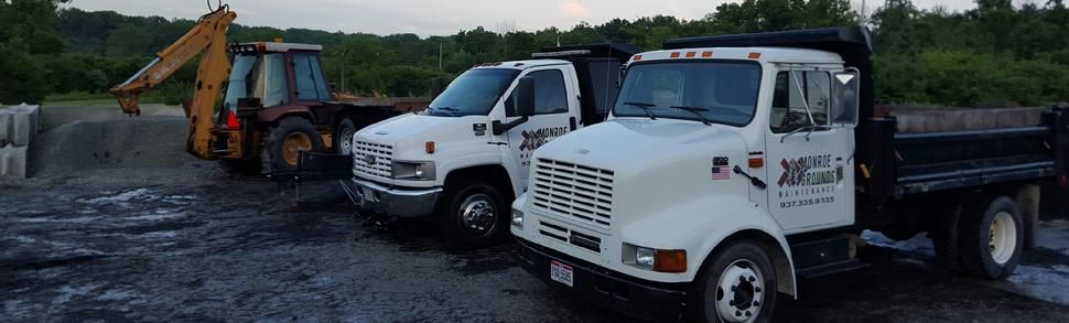 Two white dump trucks and a yellow backhoe parked on a dirt lot with a treeline in the background.