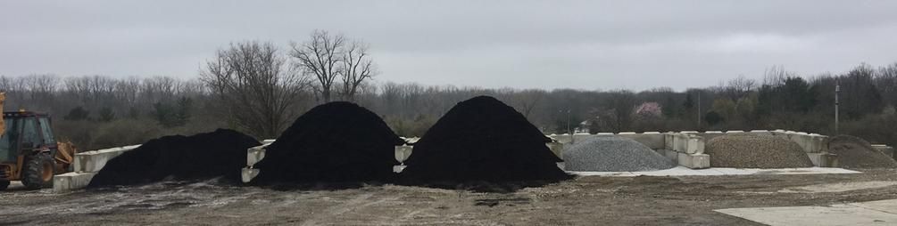 Several large mounds of black soil, gravel, and sand sit on a dirt lot with construction equipment in an overcast field.