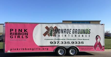 A trailer parked outdoors with branding for Pink Ribbon Girls and Monroe Grounds Maintenance, including contact details.