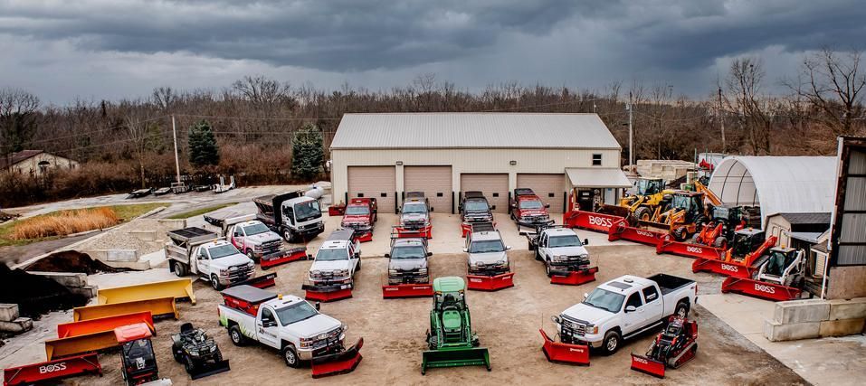 A fleet of white pickup trucks with snow plows parked in front of a service building under a cloudy sky.
