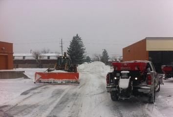 A tractor with an orange snowplow and a truck with a red salt spreader work on a snowy commercial lot.