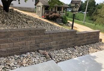 A beige brick retaining wall sits in front of a sloped, rocky hill with a small shed and birdhouse in the background.