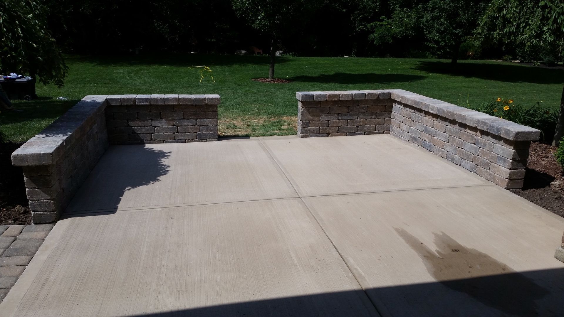 A concrete patio with a low, U-shaped stone seating wall, situated in a green backyard.