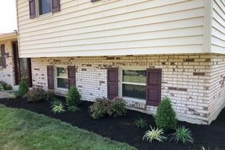 A side view of a house with beige siding, cream brick, and dark shutters, featuring a freshly mulched garden bed.