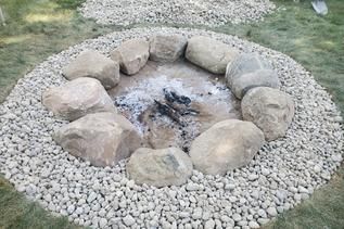 A circular fire pit surrounded by large tan stones and a wide border of light-colored gravel on a grass lawn.