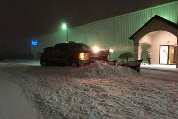 A snowplow truck clears snow in front of a commercial building entrance during a night snowfall.
