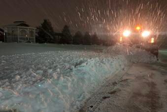 A snowplow clears a snow-covered road at night, with illuminated headlights cutting through falling snow.