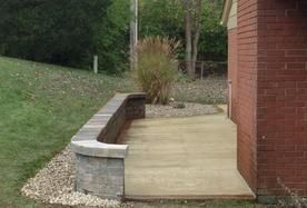 A curved grey stone retaining wall separates a concrete patio from a grassy hillside next to a red brick building.