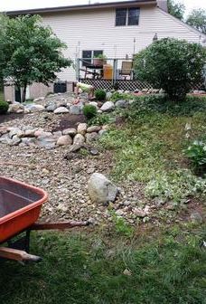 An orange wheelbarrow in the foreground of a sloped backyard with a rock garden and a multi-story house in the background.