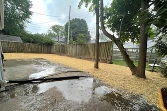 A backyard with a concrete patio showing standing water, bordered by a large area covered in fresh wood mulch and a fence.