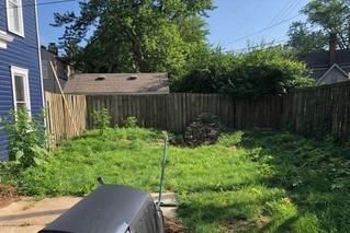 A backyard with a blue house on the left, an overgrown lawn, wooden fencing, a garage in the back, and a grill in front.