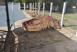 A swimming pool deck area undergoing renovation with a large pile of excavated dirt next to a white fence.