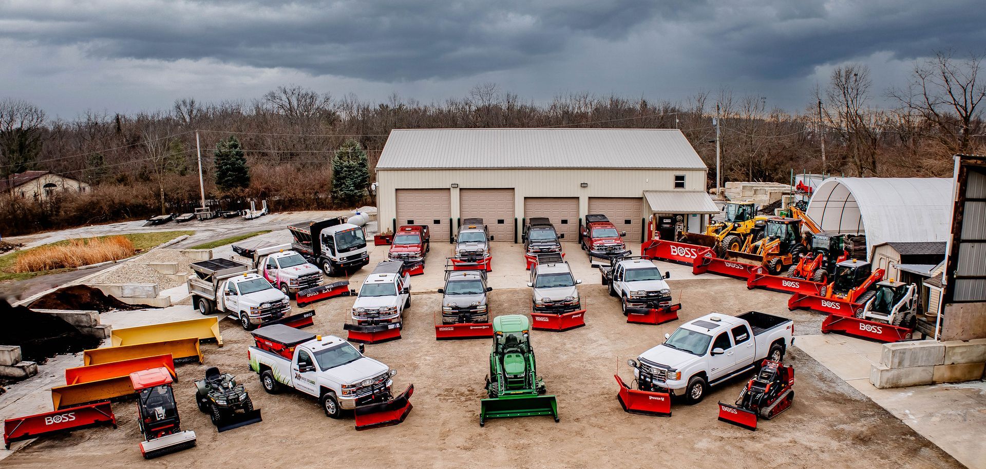 A fleet of plow-equipped trucks and construction vehicles parked in front of a service building under a cloudy sky.