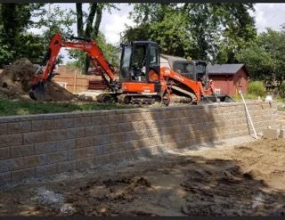 An orange excavator works behind a newly constructed stone retaining wall in a backyard.