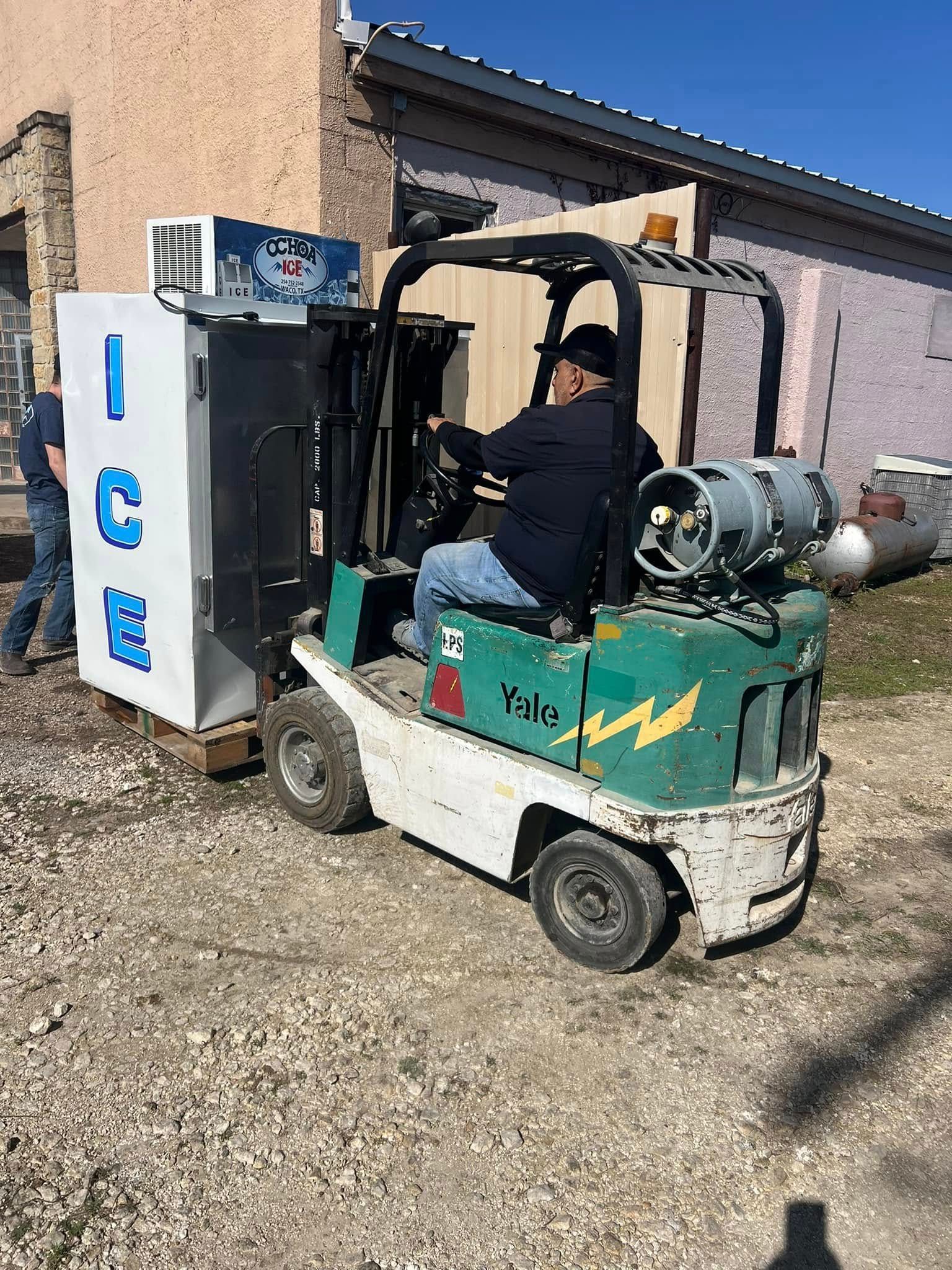 A man is driving a forklift next to a refrigerator.