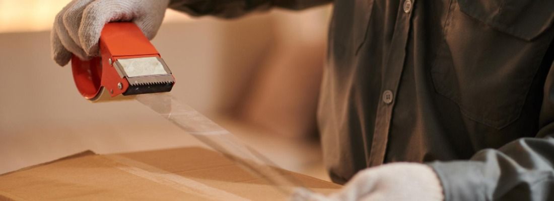 Person using a red tape dispenser to seal a cardboard box on a table