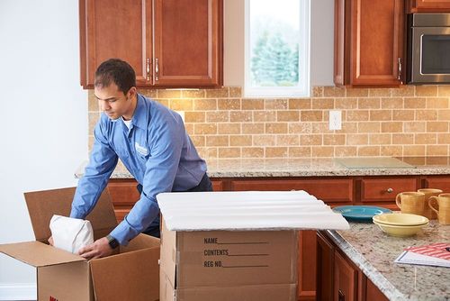 Man packing dishes into a box in a kitchen with wooden cabinets and a tiled backsplash