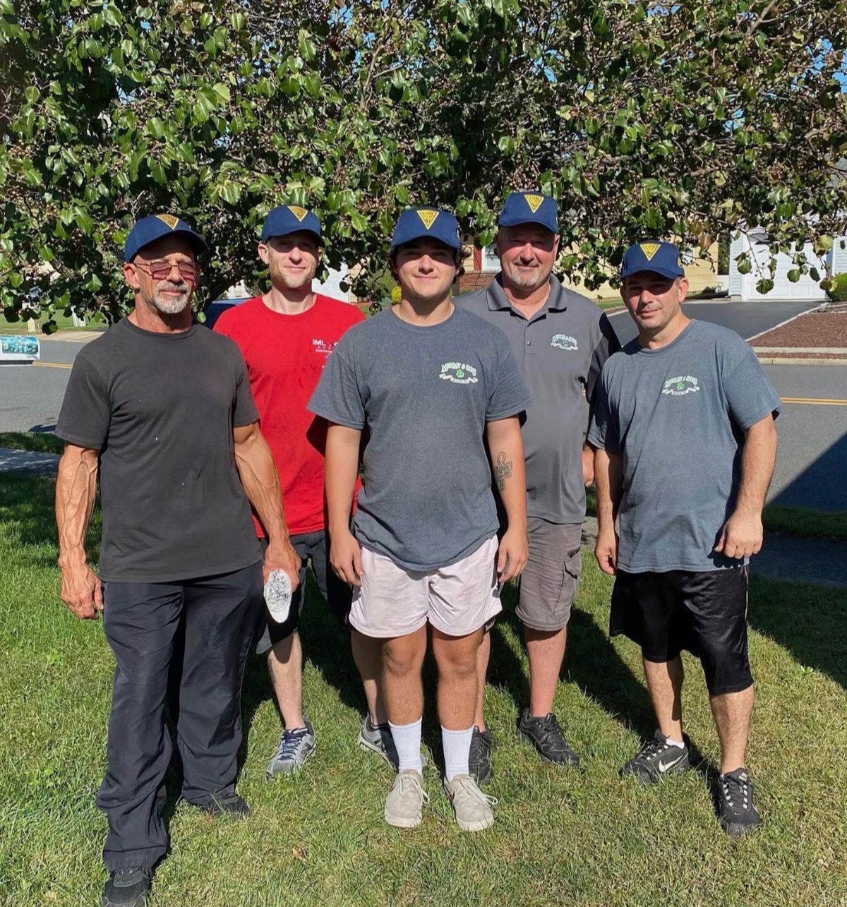 Five men in baseball caps stand together on grass outdoors near a tree and parking lot.