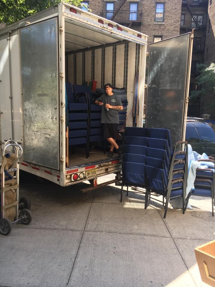 Person unloading stacked blue chairs from a truck on a city sidewalk