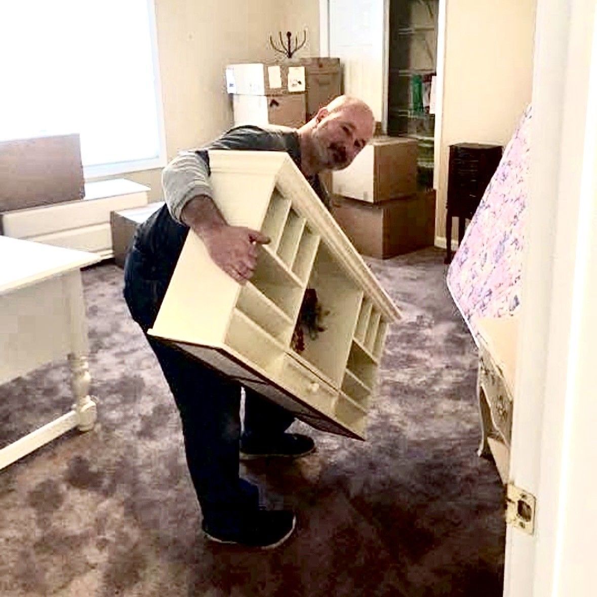Man moving a large beige shelving unit through a carpeted room with boxes in the background