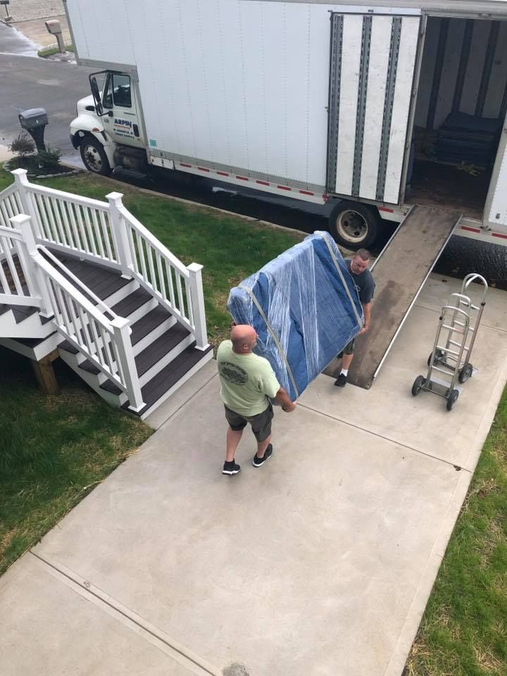 Two people move a blue-covered item down a driveway beside a white moving truck and house stairs.