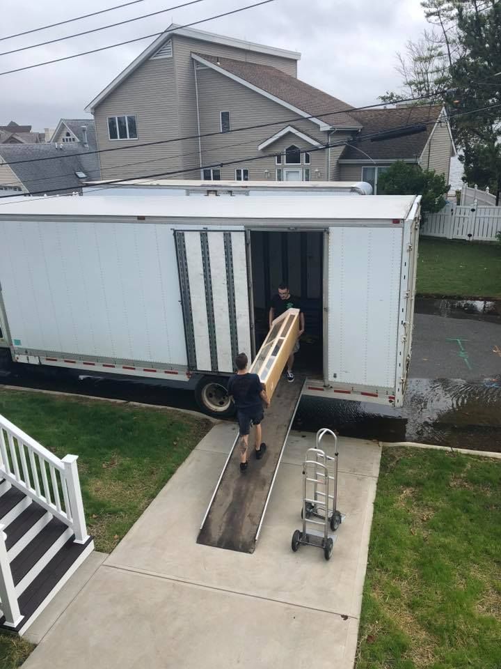 Worker unloading a large beam from a white moving truck at a house entrance, with a ramp and dolly nearby.