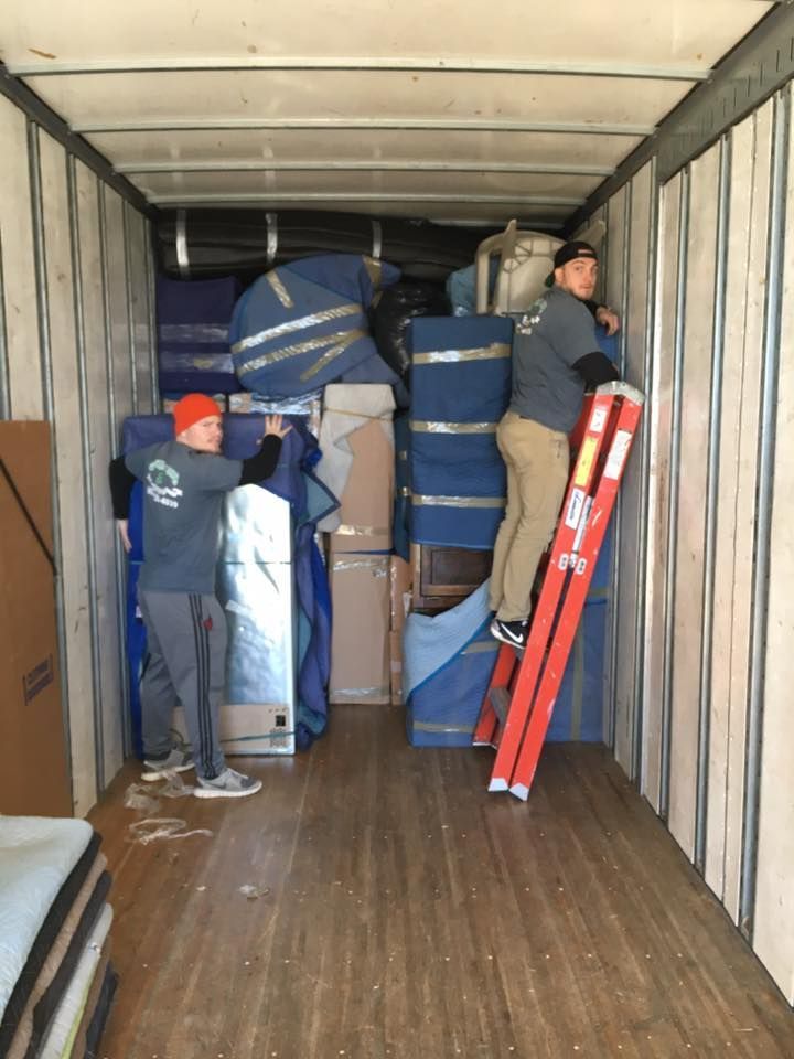 People loading wrapped furniture and boxes into a moving truck, with a red ladder leaning inside.