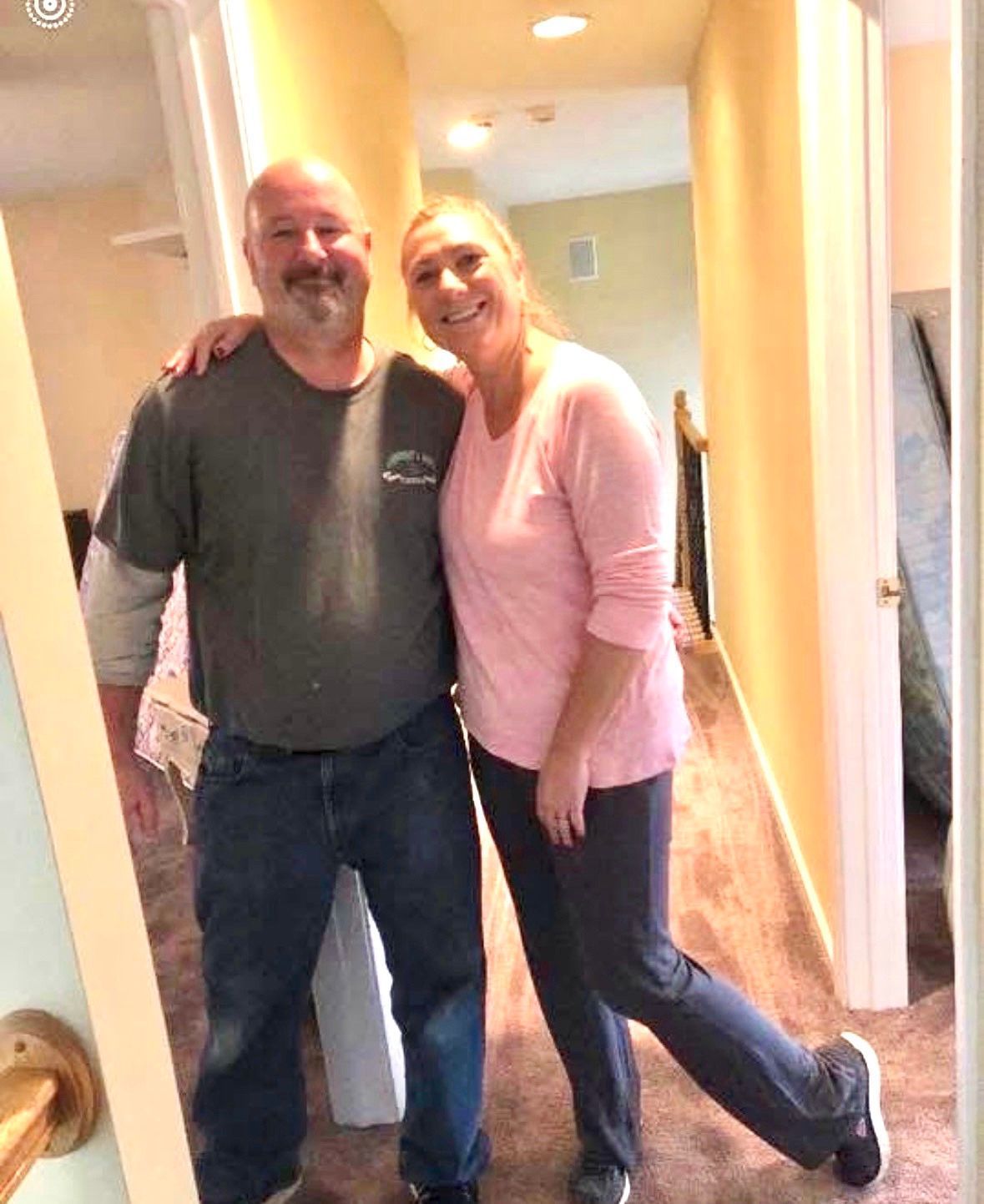 Two people smiling and posing together in a hallway, one arm around the other.