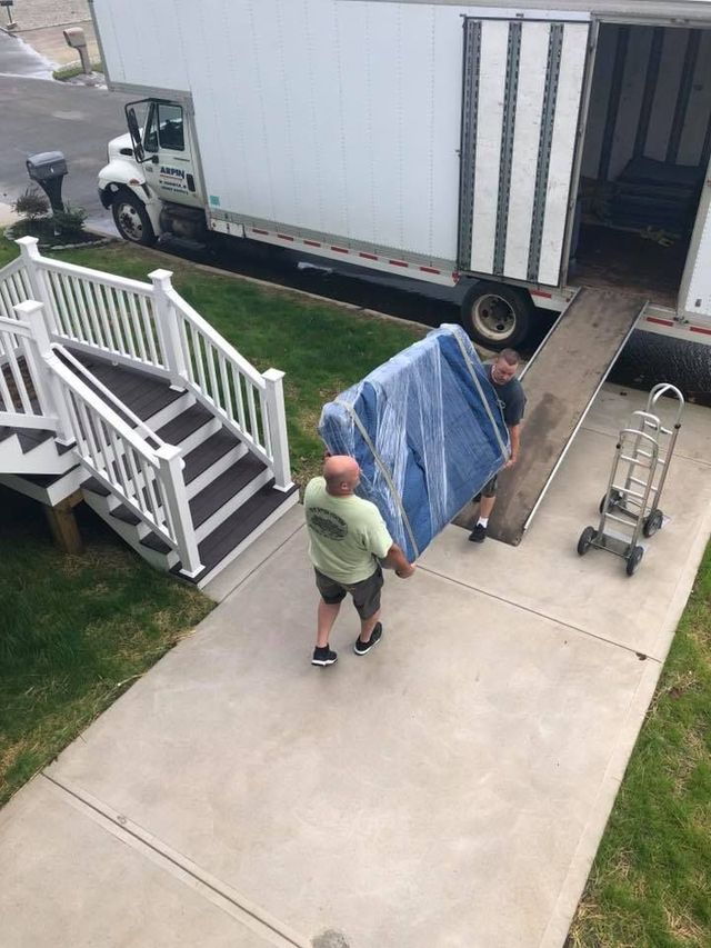 Person moving a blue-covered item on a dolly beside a moving truck and house porch stairs