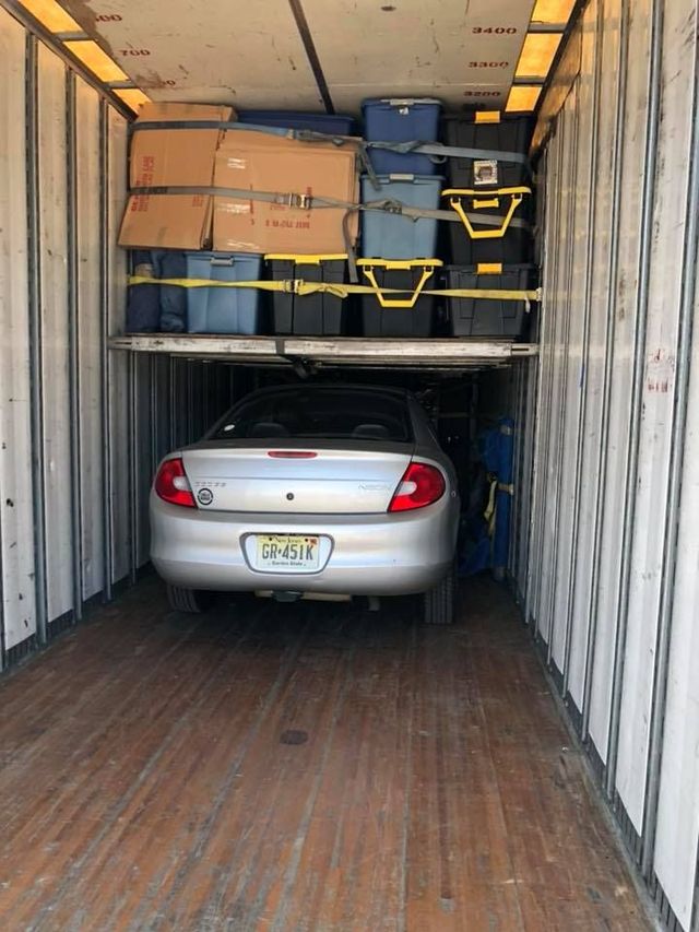 Silver car parked inside a moving truck with stacked boxes and furniture above it