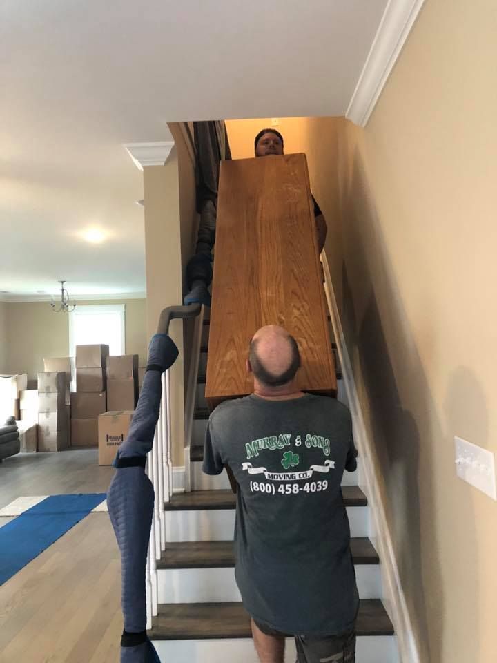 Two movers carry a large wooden cabinet up a narrow staircase in a hallway.