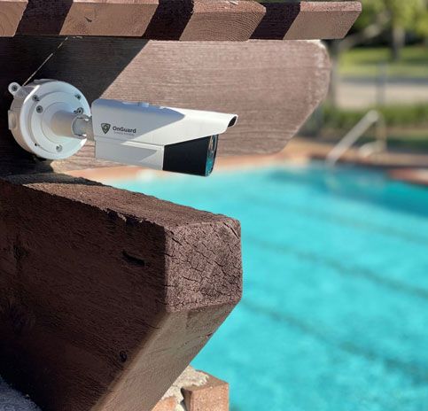 Security camera mounted on a wooden beam overlooking a blue swimming pool.