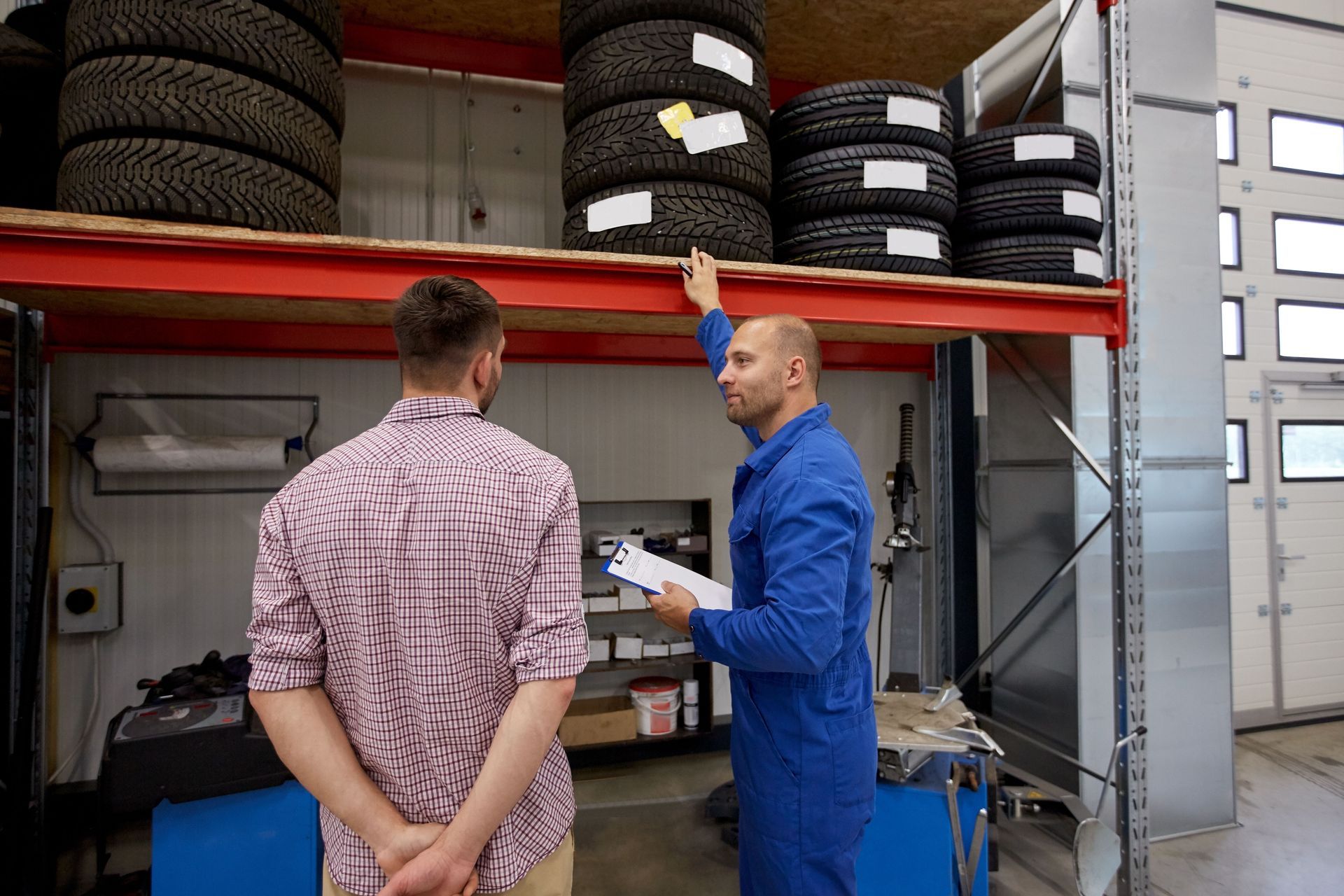 Mechanic in blue overalls points to tires on shelf, talking to a customer in a garage.