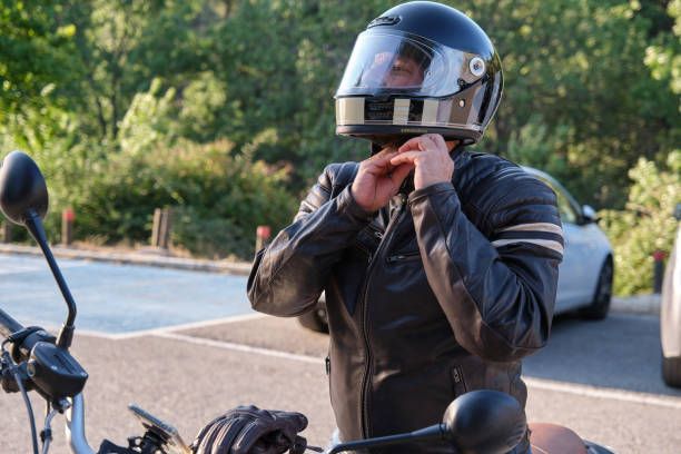 Motorcyclist adjusting helmet strap beside a parked bike on a roadside.