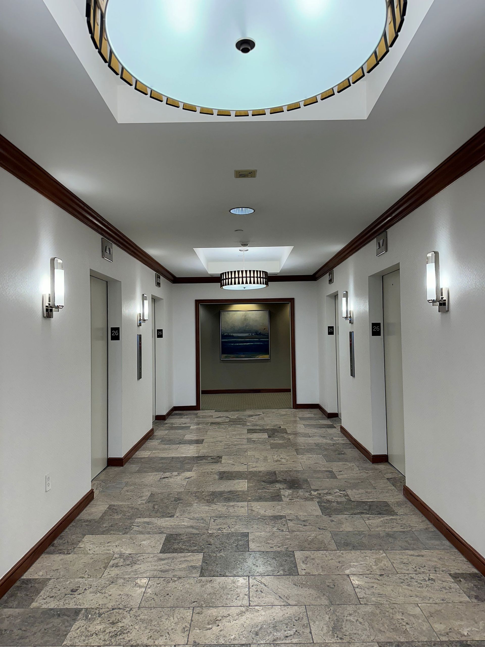 Empty hotel hallway with white walls, marble floor, wall sconces, and circular ceiling skylight