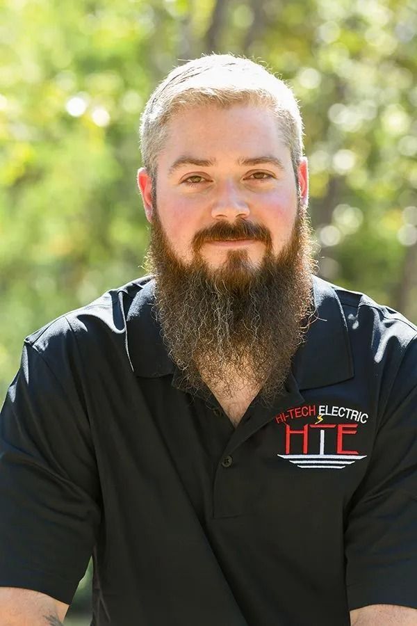 Man with a full beard wearing a black polo shirt with logo outdoors, smiling.
