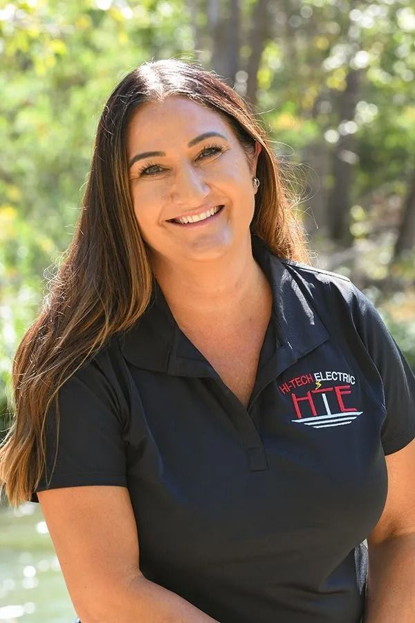 Woman with long brown hair smiles outdoors, wearing a black shirt with logo.