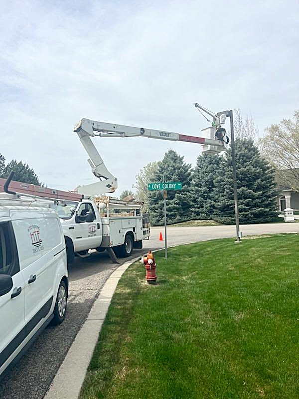 Utility truck with extended boom working on a street sign at a corner with green grass and trees.