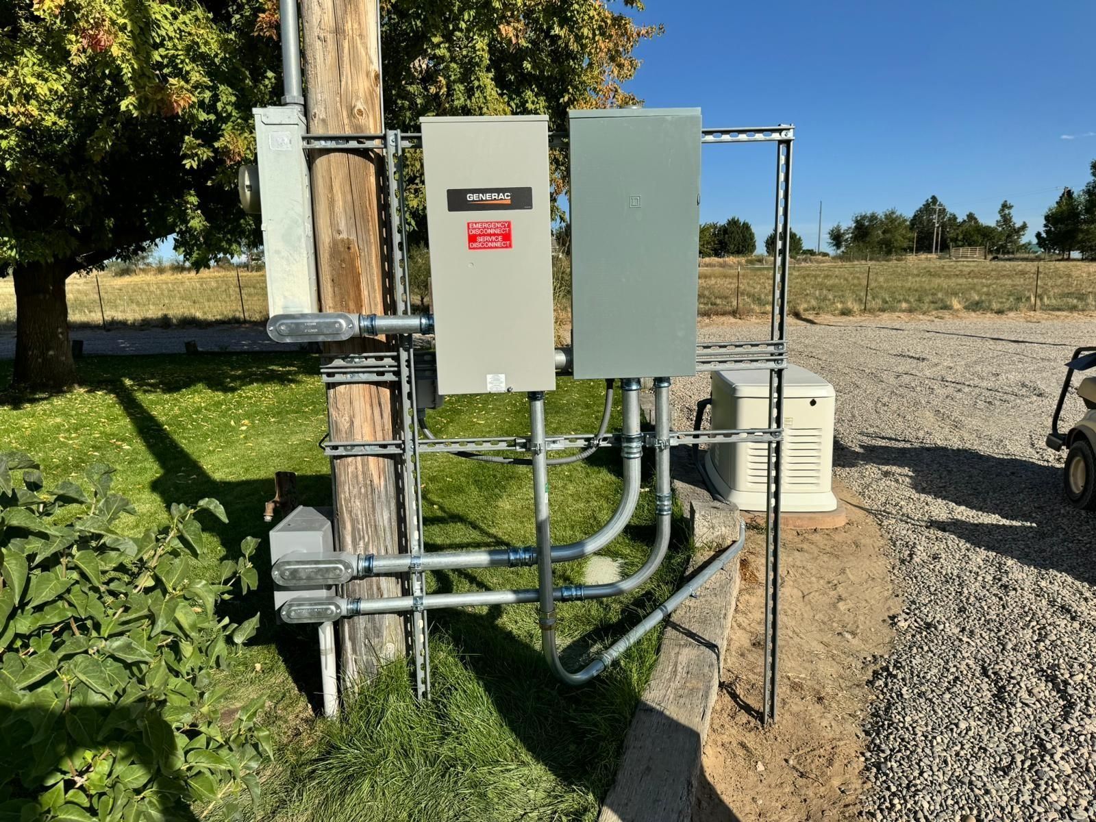 Electrical boxes and conduit mounted on a pole next to a gravel driveway and grass.